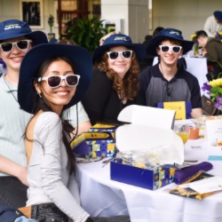 A 集团 of smiling young adults sits at a table, each wearing white sunglasses and matching blue hats. They are surrounded by gift boxes, pamphlets, and floral arrangements. The setting appears to be a casual event or gathering.